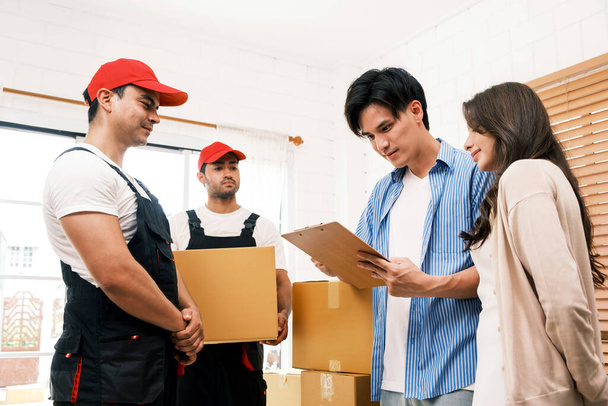 A couple signs the checklist of Asian and Caucasian workers in uniform unloading cardboard boxes from the truck. Delivery men unloading boxes to couple home. Professional delivery and moving service. - Photo, Image
