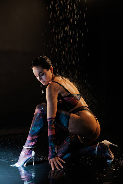 Young sexy woman poses in lingerie in BDSM style, long stockings, gloves and high heels shoes under pouring water drops against black background. - Photo, Image