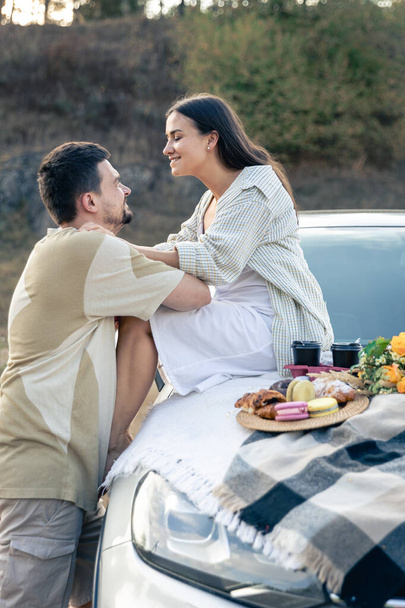 Mooi jong paar genieten van picknick tijd op de zonsondergang in een weide, leunend op ouderwetse auto, man en vrouw genieten van elkaar. - Foto, afbeelding