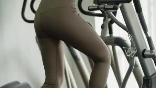 Una joven esbelta atlética pedalea una máquina de ejercicios en el gimnasio. Fácil y bastante régimen de entrenamiento. Beneficios para el cuerpo y la relajación agradable en una acción. La mujer mira a la ventana. Alto. - Metraje, vídeo