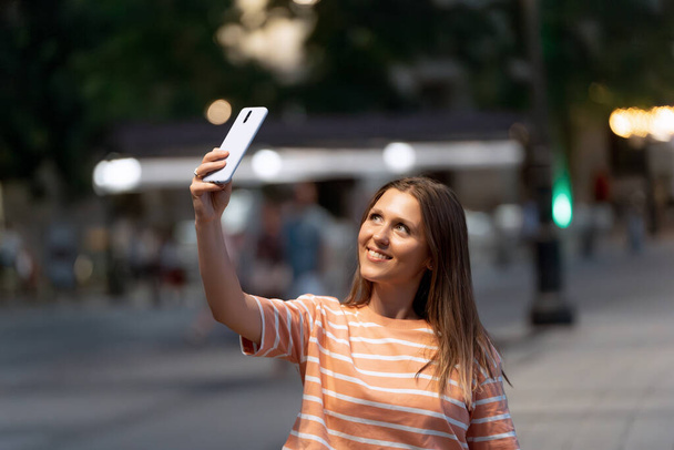 Nainen hymyilee ottaessaan selfietä matkapuhelimen kanssa ulkona kadulla yöllä. Teknologian käsite. - Valokuva, kuva