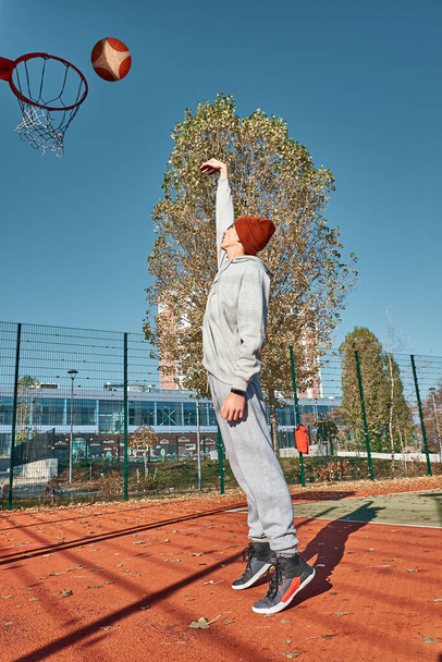 Ein Teenager in grauem Trainingsanzug und braun schießt ein Tor im Ring. Sportler auf dem Basketballplatz. Basketballspieler. Sportgeräte. - Foto, Bild