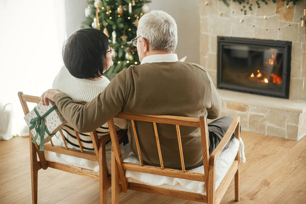 Beau couple âgé étreignant et assis à la cheminée chaude dans le salon décoré de Noël élégant. Heureuse famille aînée échangeant cadeau de Noël et profitant d'une soirée confortable. - Photo, image