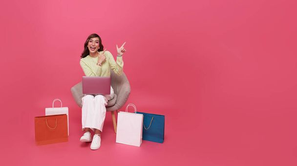 Feliz mujer asiática joven de moda sentada con la bolsa de la compra y apuntando el dedo para copiar el espacio, ella está haciendo compras en línea, Gadget con espacio libre vacío para maqueta, bandera - Foto, imagen