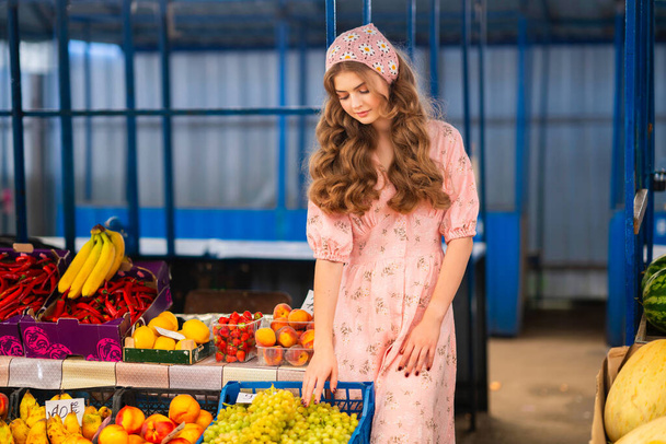 Mooi blond krullend meisje in gebreide bloemensjaal en roze jurk staan op de markt met verschillende vruchten. Jonge tiener die groenten verkoopt. - Foto, afbeelding