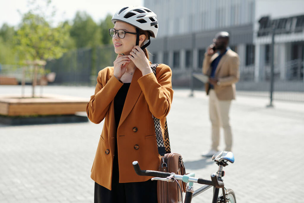 Ευτυχισμένη νεαρή επιχειρηματίας σε formalwear στερέωση ζώνη του κράνους ασφαλείας από το αυτί πριν κάθεται στο ποδήλατο και ιππασία στο κέντρο του γραφείου - Φωτογραφία, εικόνα