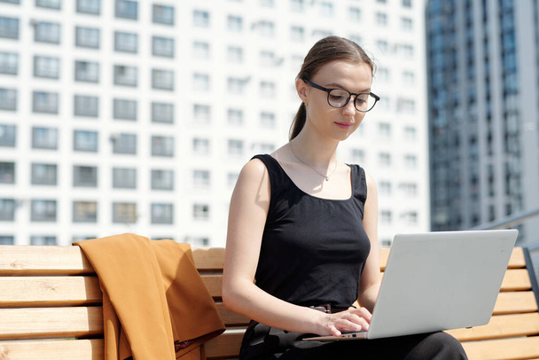 Молодая бизнесвумен в очках и черных жилетах печатает и смотрит на экран ноутбука, сидя на скамейке и организуя работу на открытом воздухе - Фото, изображение