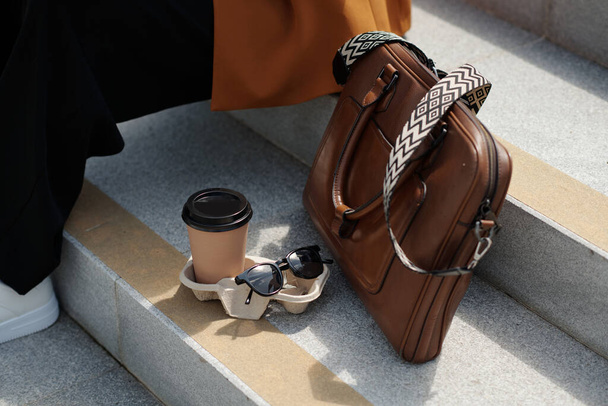 Composición de gafas de sol, taza de café y bolso de cuero marrón en escaleras de hormigón gris de escalera donde joven empleada sentada - Foto, Imagen