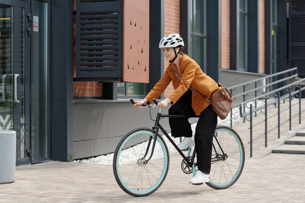 Vista lateral de la joven mujer de negocios en traje de lujo tranquilo y casco de protección montar en bicicleta a lo largo de edificio moderno de centro de oficina después del trabajo - Foto, Imagen