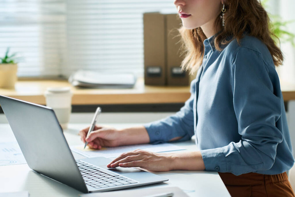 Side view of young serious female white collar worker in blue shirt sitting by desk in front of laptop, making notes and analyzing online data - Foto, Imagen