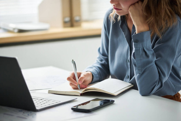 Przycięte zdjęcie młodej bizneswoman lub studentki w niebieskiej koszuli trzymającej długopis nad otwartym notebookiem, myśląc jednocześnie o nowych pomysłach na pracę w biurze - Zdjęcie, obraz