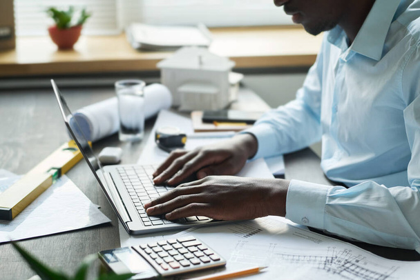 Вид сбоку молодого архитектора-мужчины, сидящего за письменным столом с чернилами, ручными инструментами, калькулятором и другими вещами и сидящего на клавиатуре ноутбука - Фото, изображение