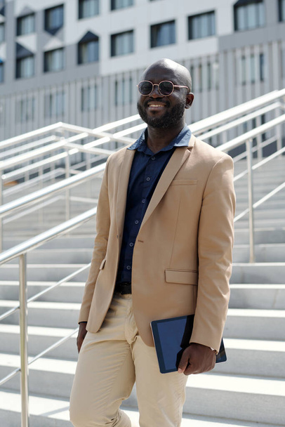 Jeune solpreneur afro-américain à succès en tenue de luxe silencieuse et lunettes regardant la caméra tout en se tenant debout contre l'escalier - Photo, image