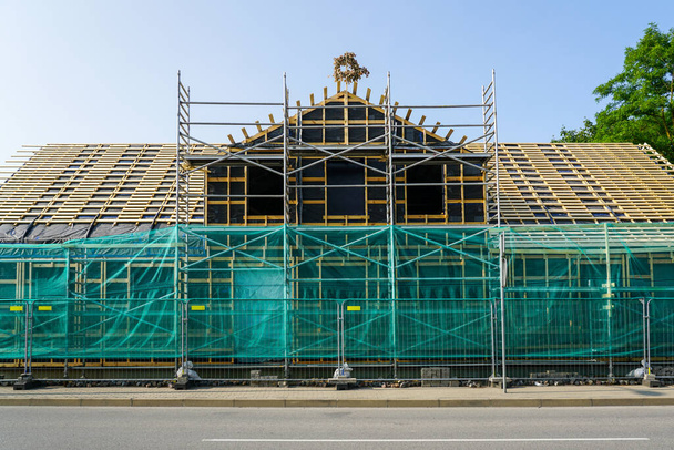 Vista frontal de la fachada de una casa de madera en restauración con andamios y red de seguridad, nuevo torno de techo, valla metálica temporal - Foto, Imagen