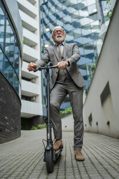 один мужчина старший кавказский мужчина, стоящий на электрическом мотороллере езда в городе электронный скутер экологически чистый способ транспорта реальные люди бизнесмен носить костюм перед копировальной площади компании - Фото, изображение
