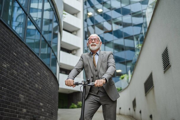один мужчина старший кавказский мужчина, стоящий на электрическом мотороллере езда в городе электронный скутер экологически чистый способ транспорта реальные люди бизнесмен носить костюм перед копировальной площади компании - Фото, изображение
