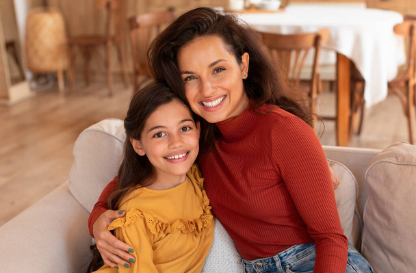 Mothers Love. Portrait Of Young Woman Hugging Her Cute Daughter While Resting On Couch At Home Interior, Loving Mom Bonding With Kid Enjoying Time Together Sitting On Sofa Indoor - Photo, Image