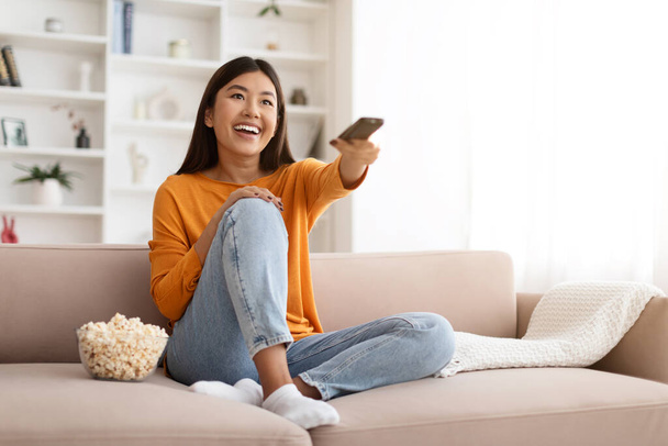 Счастливая молодая дама проводит свое свободное время дома, смотрит телевизор и ест кукурузу на диване, свободном пространстве. Волнующая китаянка с пультом дистанционного управления наслаждается интересным фильмом - Фото, изображение