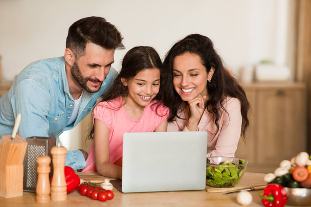 Szczęśliwa rodzina trzech Europejczyków korzystających z laptopa, przeglądanie zdrowych przepisów podczas przygotowywania kolacji razem, wnętrze kuchni. Rodzice i gotowanie córki - Zdjęcie, obraz