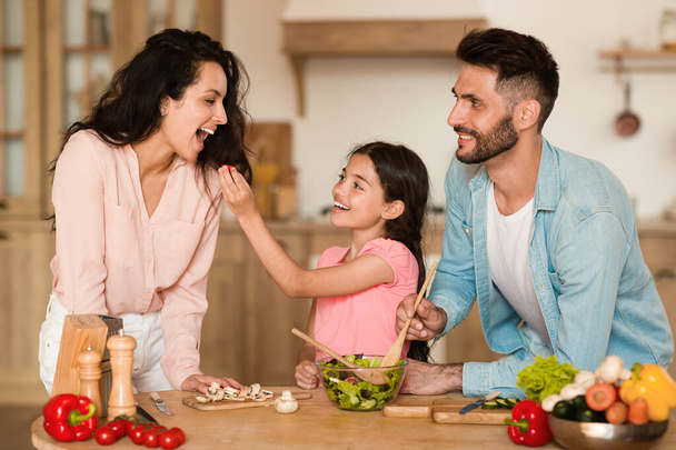 Веселая дочь кормит свою счастливую мать маленьким помидором черри во время приготовления салата вместе с родителями. Позитивные мама, папа и девочка готовят за столом на кухне - Фото, изображение