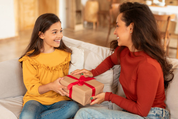 Smiling Kid Girl Giving Birthday Gift To Mom Sitting Together On Sofa, Celebrating Family Holiday At Home. Mothers Day Celebration, Surprises And Presents Offer Concept. Selective Focus - Foto, imagen