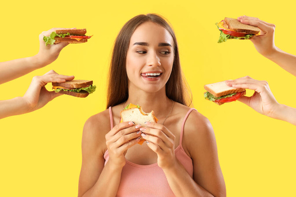 Όμορφη νεαρή γυναίκα που περιβάλλεται από χέρια με νόστιμα σάντουιτς σε κίτρινο φόντο - Φωτογραφία, εικόνα