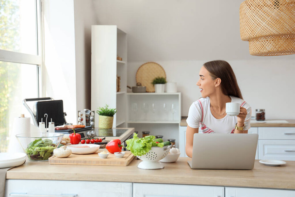 Mulher bonita usando laptop enquanto cozinha deliciosas salsichas na moderna churrasqueira elétrica na cozinha - Foto, Imagem