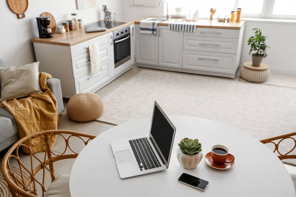 Kannettava tietokone matkapuhelimella ja kahvikuppi ruokapöydällä keittiössä - Valokuva, kuva