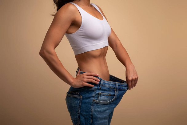 Conceito de dieta de perda de peso bem sucedida. Tiro cortado de jovem senhora em jeans grandes demonstrando resultados de seu programa de emagrecimento, promovendo a alimentação saudável, vista lateral, close-up, fundo bege - Foto, Imagem
