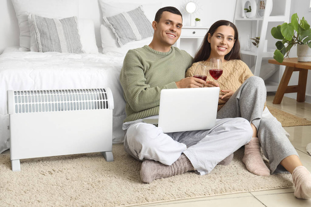 Young couple with wine and laptop warming near radiator in bedroom - Photo, Image