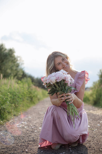 Joyeux blond charmant avec bouquet de pivoines roses douces sourit coquettiquement le jour de l'été sur la route de campagne. Fémininité et beauté. Des émotions positives. Humeur sereine. Anniversaire, cadeau. Des fleurs pour vous.  - Photo, image