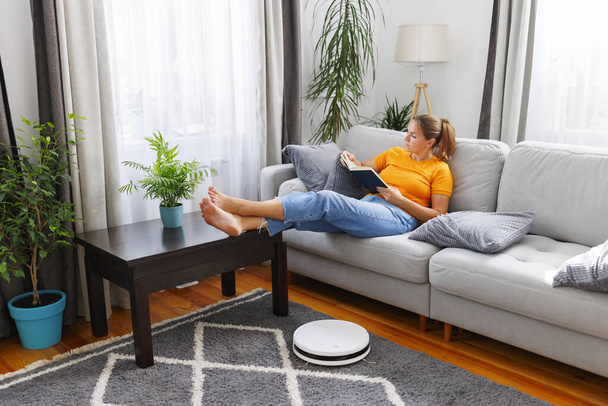 Роботизированный пылесос убирает комнату, в то время как женщина отдыхает, читая книгу на диване. Инновационные технологии очистки - Фото, изображение