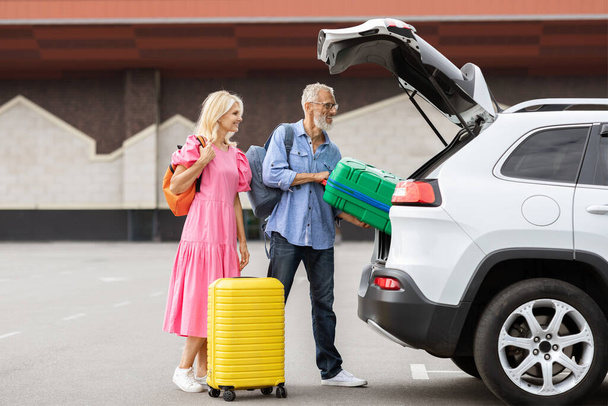 Happy caucasian senior couple going car trip together. Cheerful elderly man and woman in casual travelers getting ready before journey by auto, loading vehicle trunk with luggage, stand outdoors - Photo, Image