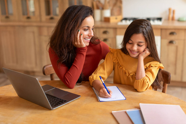 Szczęśliwa mamusia przytula swoją mądrą córkę uczennicę podczas pisania pracy domowej w pobliżu nowoczesnego laptopa wewnątrz, matka i dziecko pracy na szkolnym projekcie siedzi razem w domu - Zdjęcie, obraz
