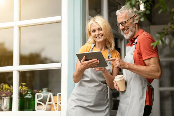Tecnologias modernas. Smiling maduro agricultores casal em aventais usando Digital Tablet juntos e beber café enquanto estiver em pé no terraço perto da casa de campo, felizes cônjuges navegando na Internet em Gadget - Foto, Imagem