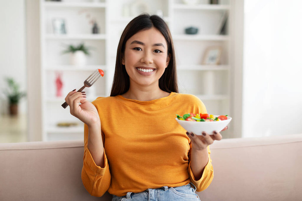 Joyeux attrayant mince jeune femme asiatique assise sur le canapé à la maison, manger de la salade faite de légumes biologiques frais, déjeuner sain, copier l'espace. Mode de vie sain, régime alimentaire, concept nutritionnel - Photo, image