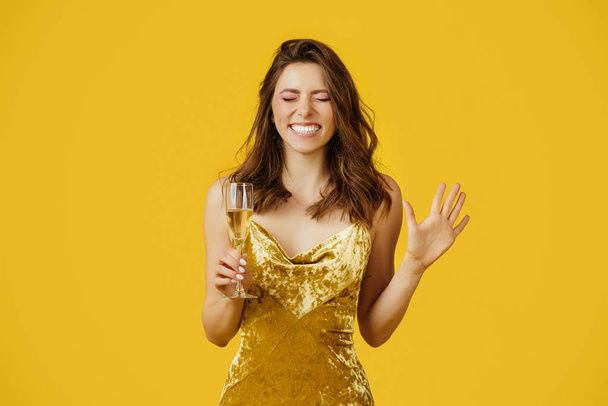 Удовлетворенная молодая европейская женщина с закрытыми глазами держит бокал шампанского и загадывает желание на желтом студийном фоне. Мечта, праздник и эмоции людей - Фото, изображение