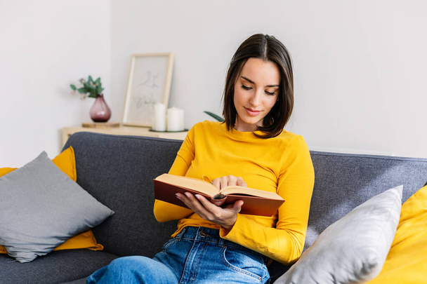 Uvolněná mladá dospělá žena čtení knihy sedí na gauči u ní doma. - Fotografie, Obrázek