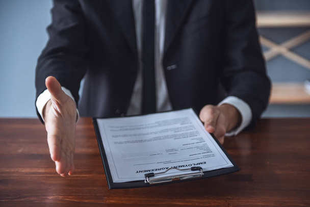 Bijgesneden afbeelding van knappe werkgever biedt zijn hand en houden een arbeidsovereenkomst zittend aan tafel in zijn kantoor - Foto, afbeelding