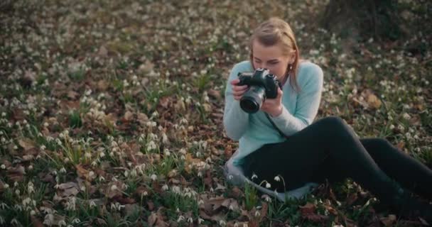 Mulher jovem loira bonita fotografando flores de neve enquanto sentado no jardim - Filmagem, Vídeo