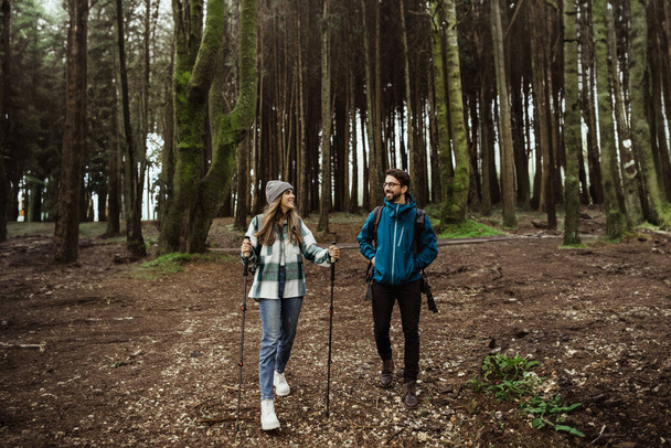 Positive young caucasian family in jackets with trekking sticks enjoy adventure, free time at weekend, walk in cold forest in autumn. Vacation together, active lifestyle, full length - Photo, Image
