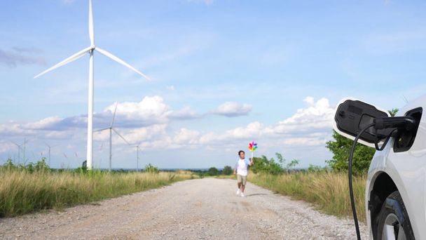 Progressif jeune garçon jouant jouet moulin à vent à côté de voiture EV au parc d'éoliennes. Générateur électrique à partir du vent par éolienne sur le côté campagne. - Photo, image