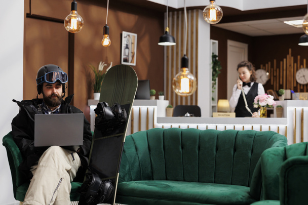 Mężczyzna snowboardzista siedzi w hotelowym lobby z wykorzystaniem Internetu na laptopie do kontroli rezerwacji. Kaukaski mężczyzna noszący odzież śnieżną za pomocą minikomputera w ośrodku narciarskim personel odpowiada telefon w tle. - Zdjęcie, obraz