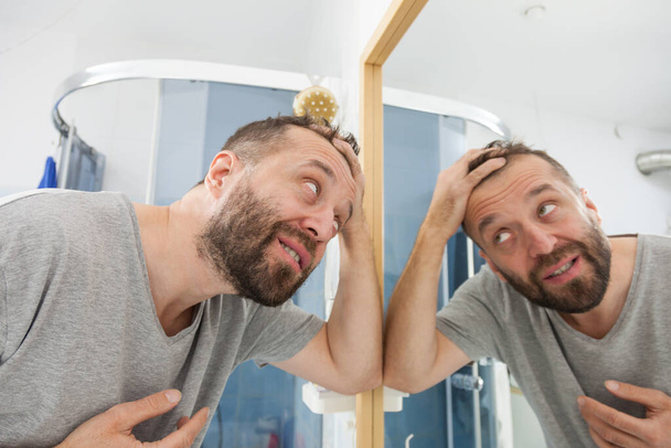 Hombre adulto preocupado mirando en el espejo del baño en su línea de cabello decreciente pensando en el tratamiento del cuidado del cabello - Foto, imagen
