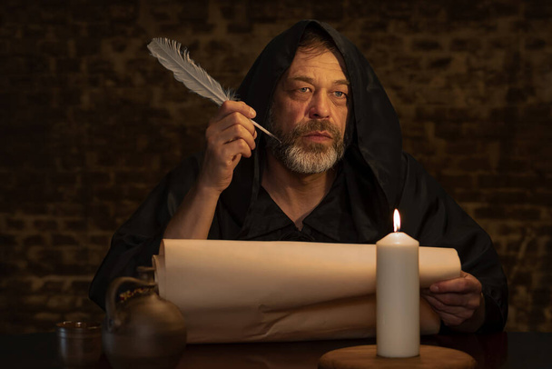 Ein alter Mönch mit Bart hält einen Federkiel in der Hand über einem Pergament vor einer dunklen Ziegelwand, dunkle Tonalität. - Foto, Bild