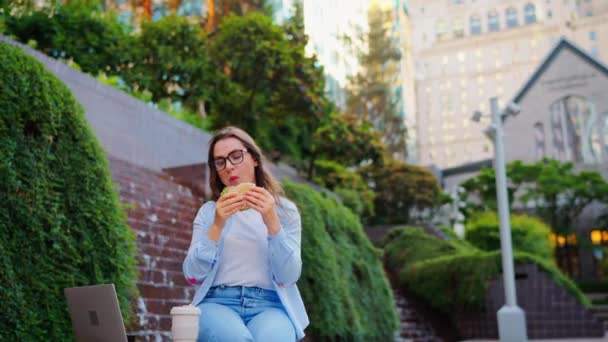 Mujer caucásica almorzando con un sándwich y café mientras está sentada en el parque con un portátil. En cámara lenta. Al aire libre. La freelancer trabaja con su portátil - Metraje, vídeo