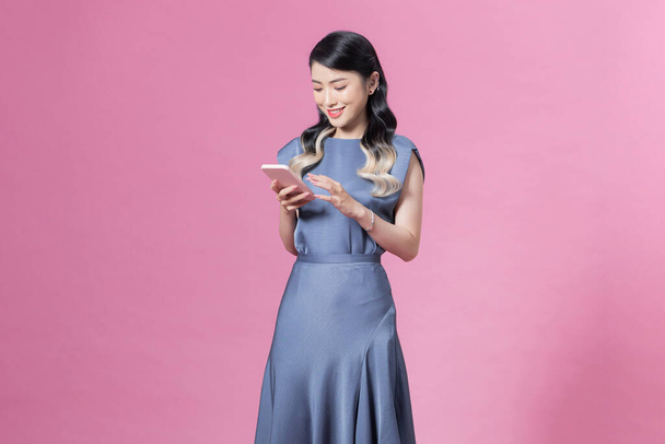 junge Frau trägt blaues Seidenkleid mit Smartphone vor rosa Hintergrund - Foto, Bild