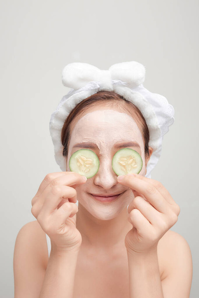 Beautiful woman with clay mask holding pieces of cucumber on white background - Photo, Image