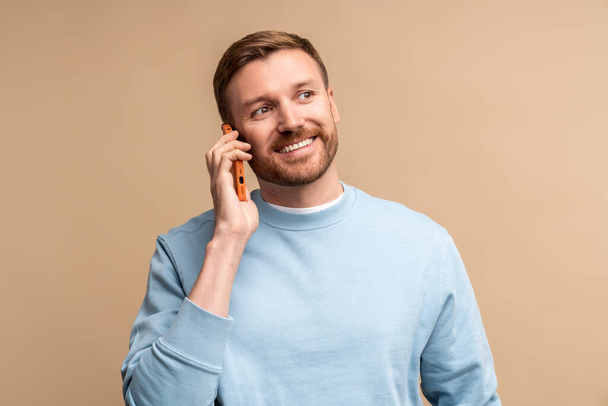 Heureux homme d'âge moyen souriant parlant téléphone sur fond beige regardant de côté. Guy profitant d'une agréable conversation sur smartphone. Portrait rire caucasien mâle communiquer sur le téléphone. - Photo, image