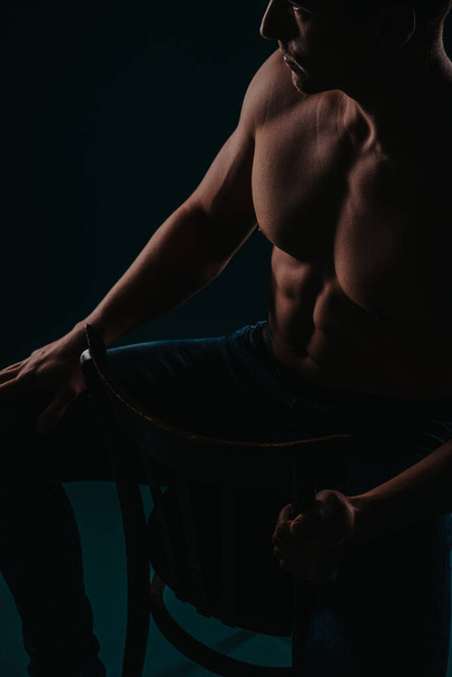 Silhouet foto van jonge, shirtloze man die zijn goed getrainde lichaam laat zien terwijl hij op een stoel zit in de studio - Foto, afbeelding